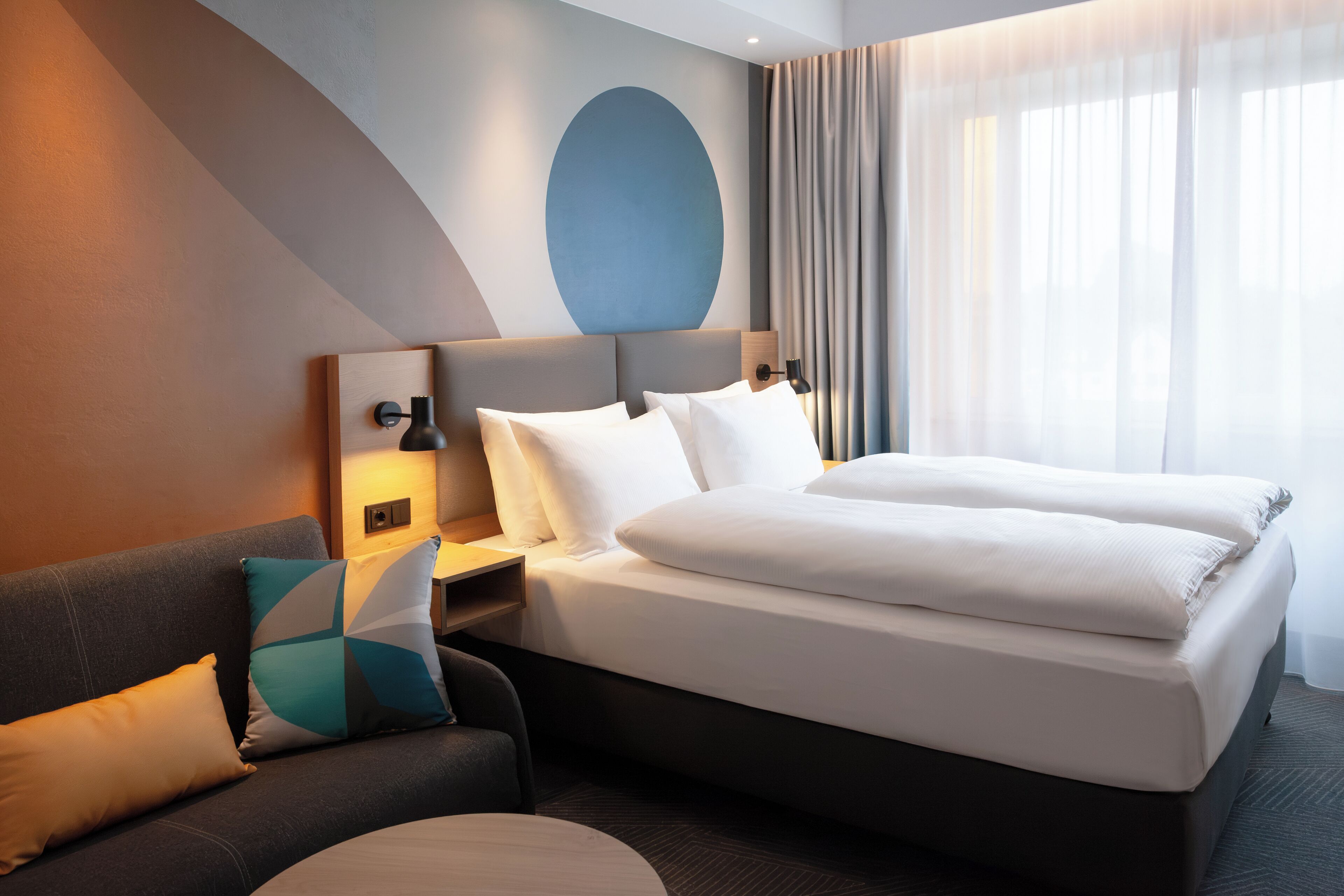 A hotel room with a bed, a sofa, a nightstand, and a wall with a geometric design.