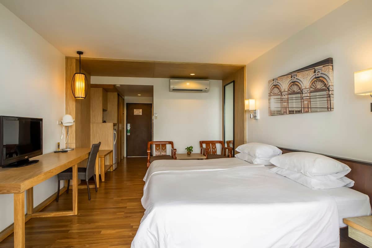 Una habitación de hotel con una cama, un escritorio con televisor, una silla, una mesita con una planta y un aire acondicionado instalado en la pared.