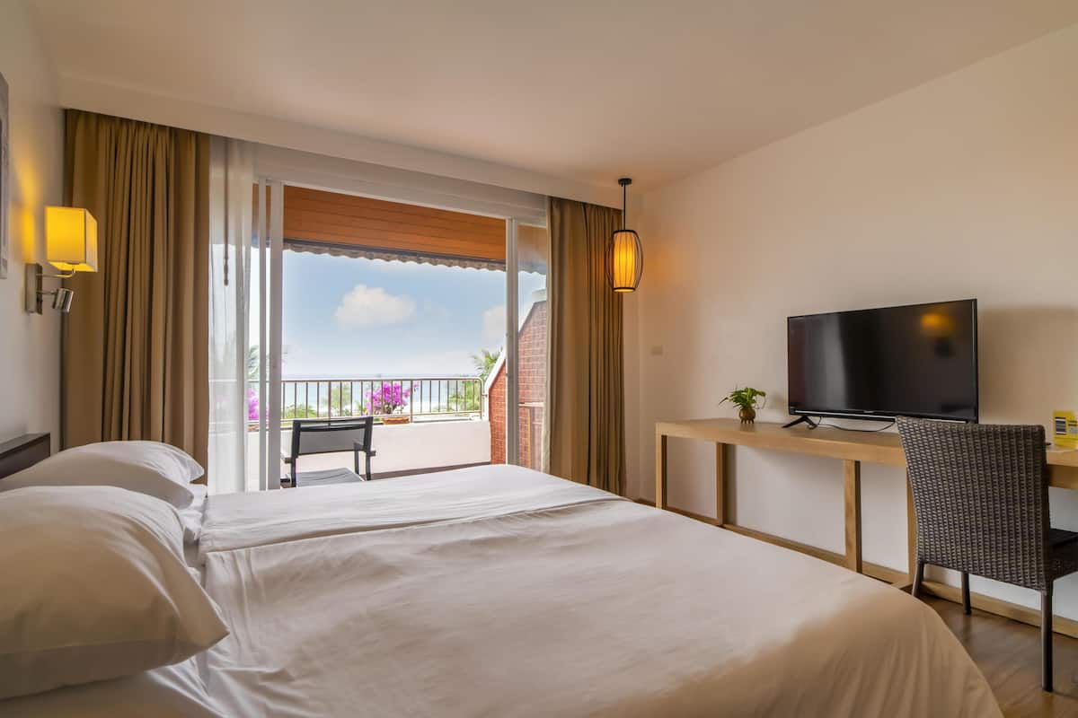 Habitación de hotel con cama, escritorio con televisor, silla y balcón con vistas.