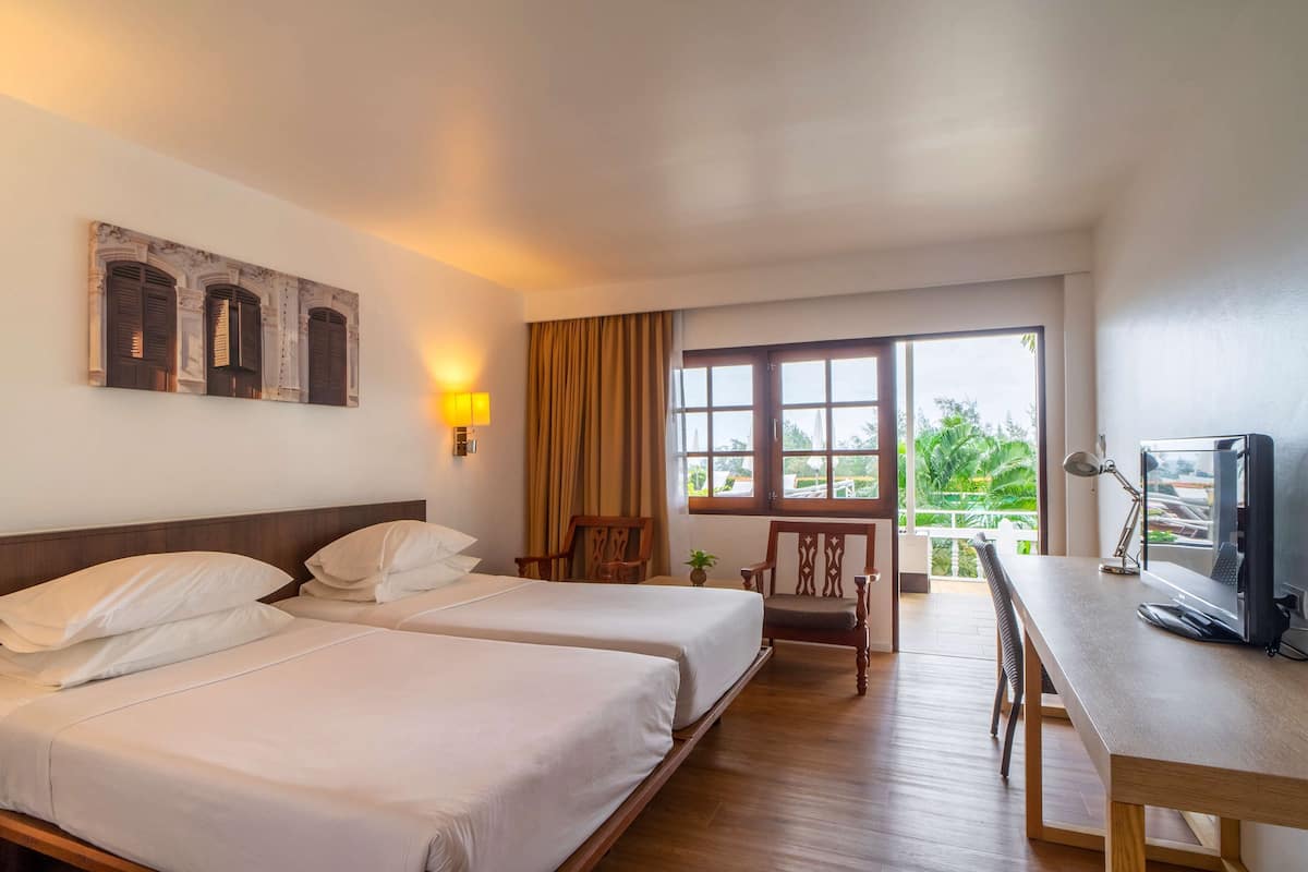 Habitación de hotel con dos camas, un escritorio, una silla, un televisor y una ventana con vistas a zonas verdes.