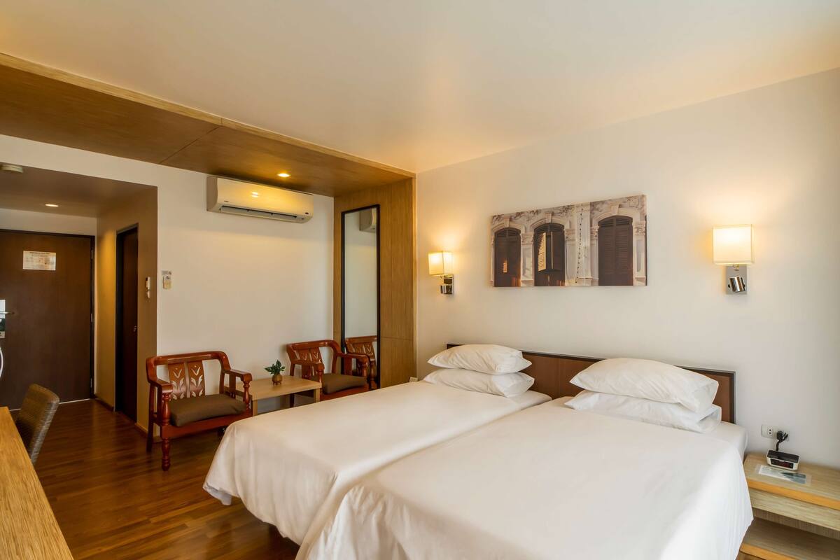 Habitación de hotel con dos camas, un banco de madera, un escritorio y aire acondicionado instalado en la pared.