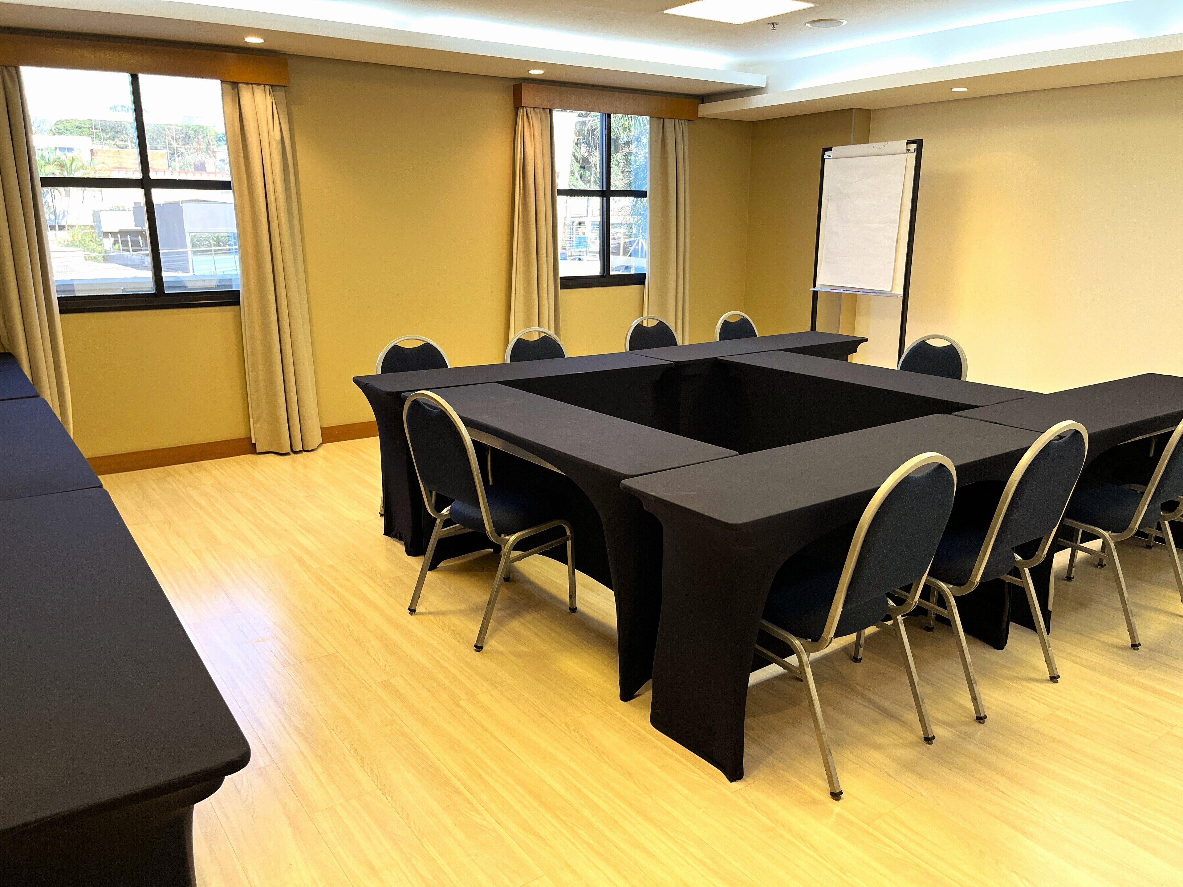 A conference room with a large rectangular table, six chairs, a whiteboard, and windows with curtains.