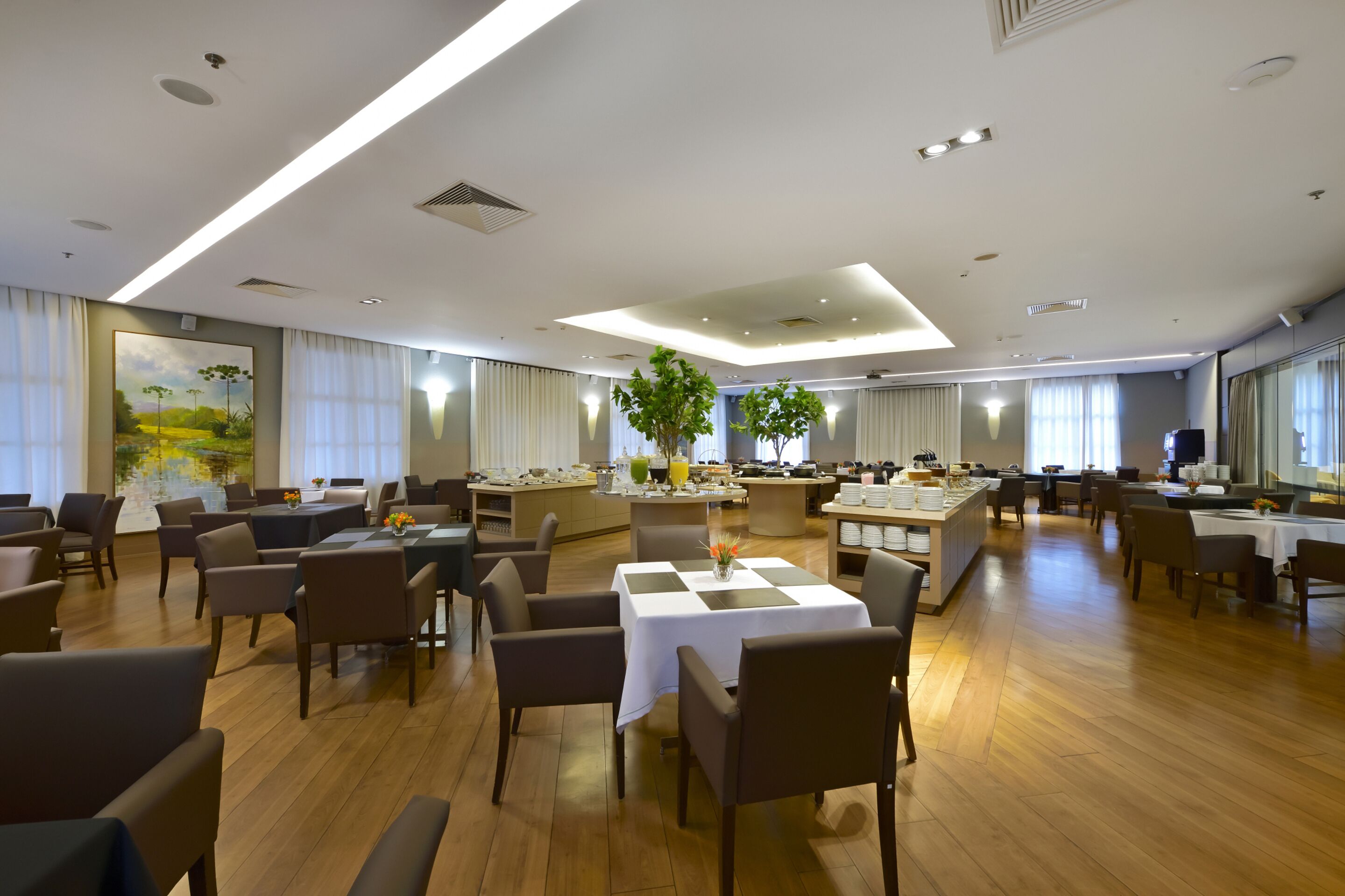 A spacious hotel dining area with multiple tables and chairs, a central island with a plant, and a large painting on the wall.