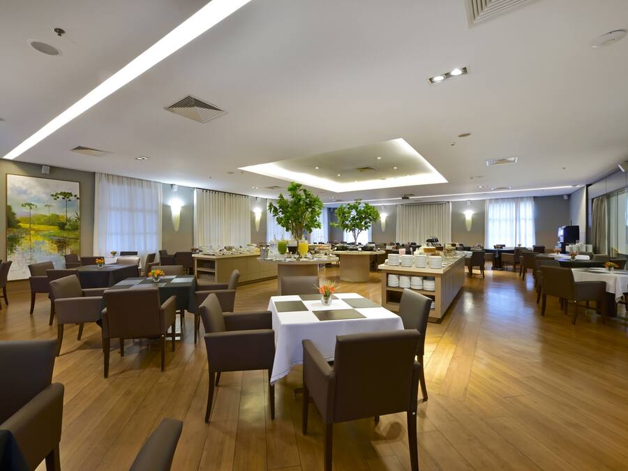 A spacious hotel dining area with multiple tables and chairs, a central island with a plant, and a large painting on the wall.
