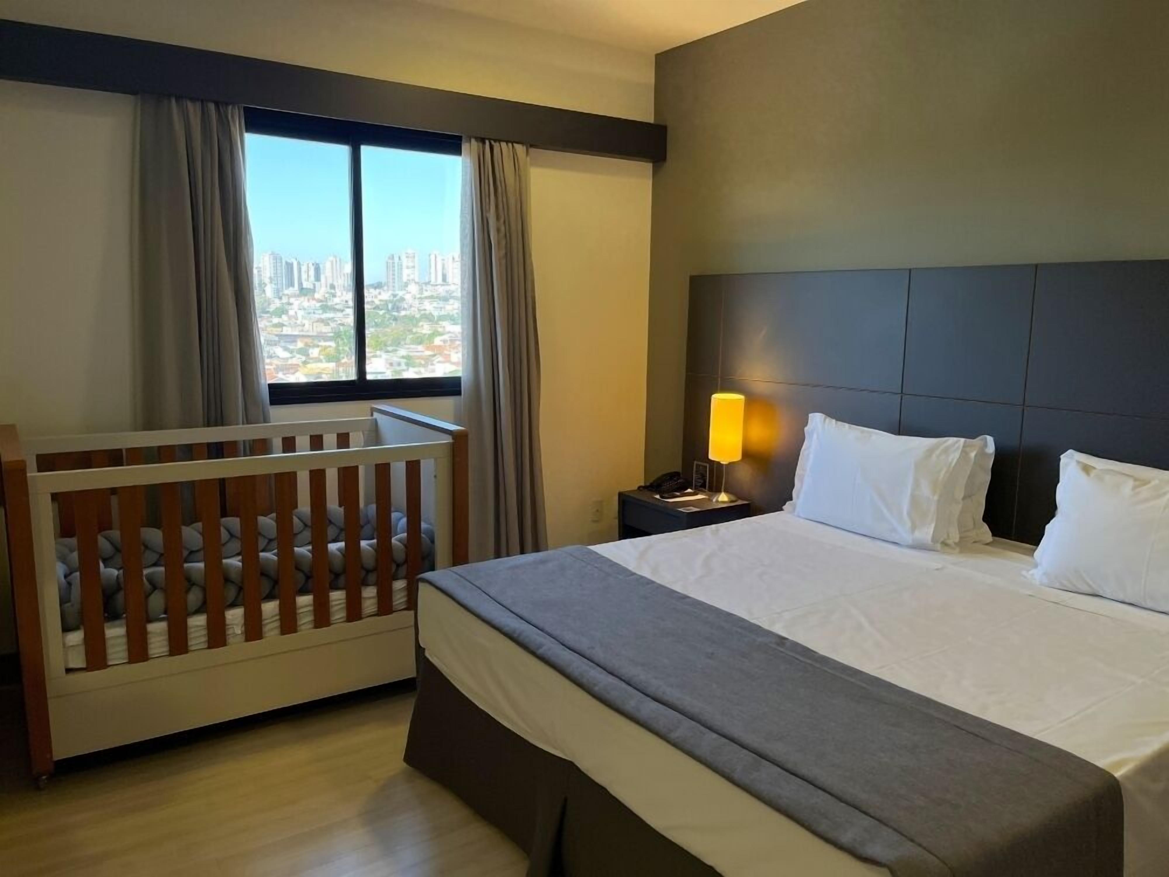Habitación de hotel con una cama grande, una cuna de madera, una mesita de noche con lámpara y vistas a un paisaje urbano a través del ventanal.