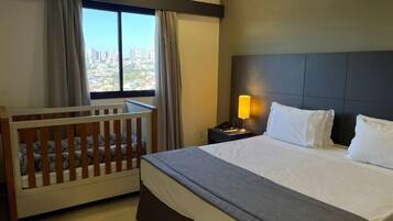 Habitación de hotel con una cama grande, una cuna de madera, una mesita de noche con lámpara y vistas a un paisaje urbano a través del ventanal.