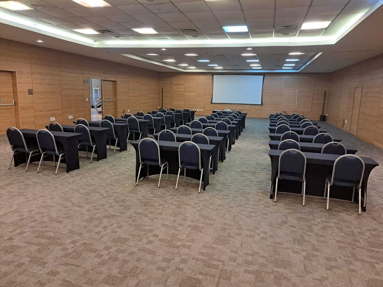 A conference room with rows of tables and chairs, a projector screen, and a carpeted floor.