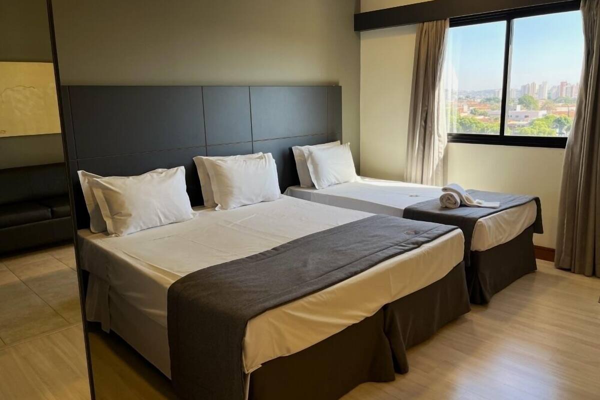 Een hotelkamer met twee bedden, een donker hoofdeinde, een groot raam met uitzicht op de stad en een houten vloer.