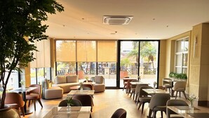 Amplia zona de estar interior con mesas, sillas, ventanas grandes y una planta en maceta.