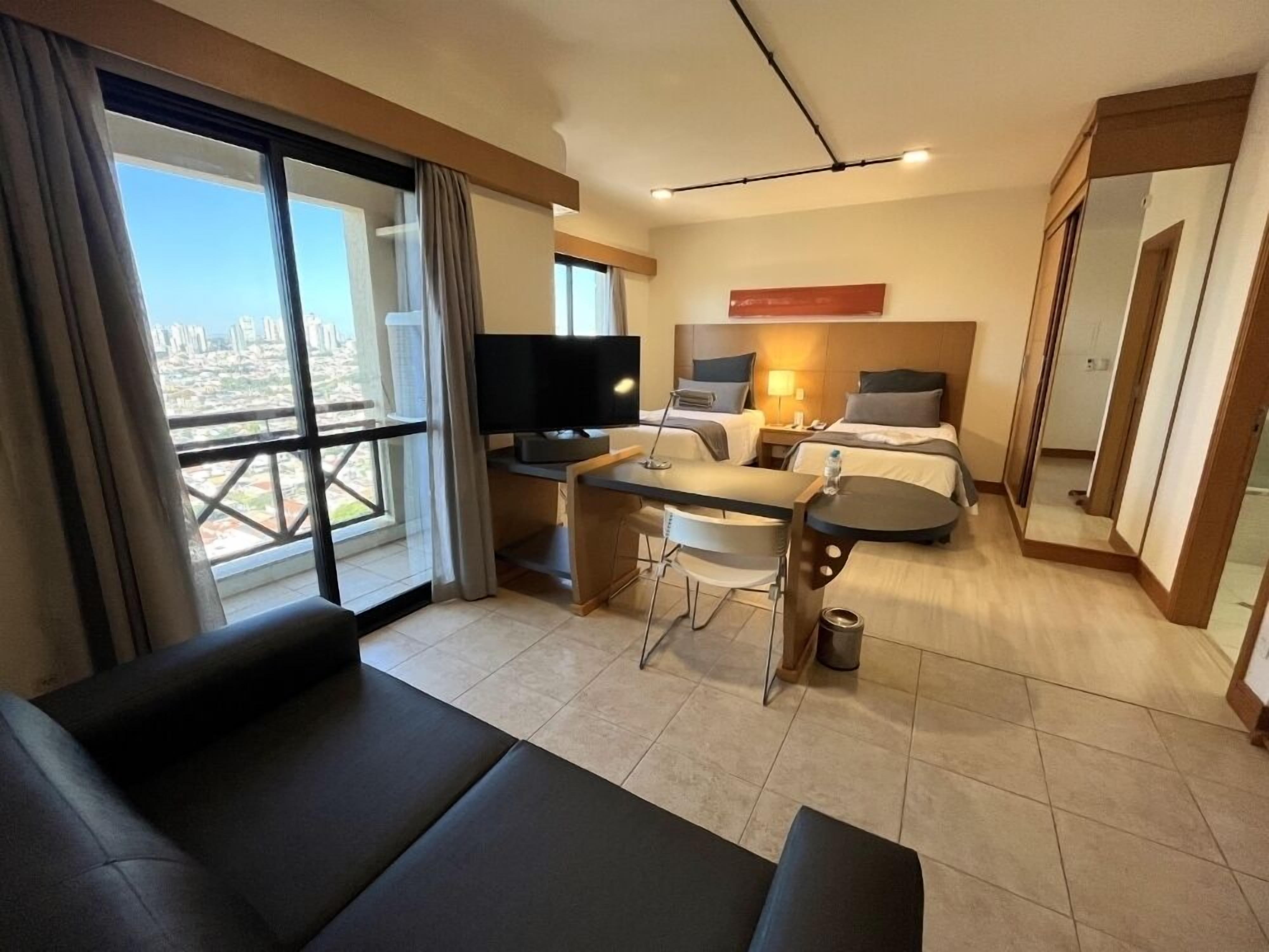 Een hotelkamer met een bank, een eettafel, een televisie, twee bedden en uitzicht op de stad.