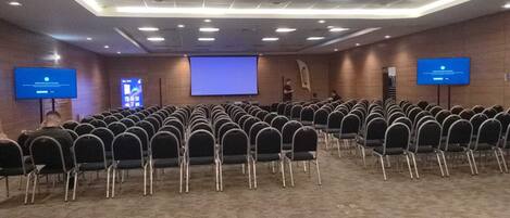 Un salón de conferencias con filas de sillas, una pantalla grande y dos monitores sobre soportes.