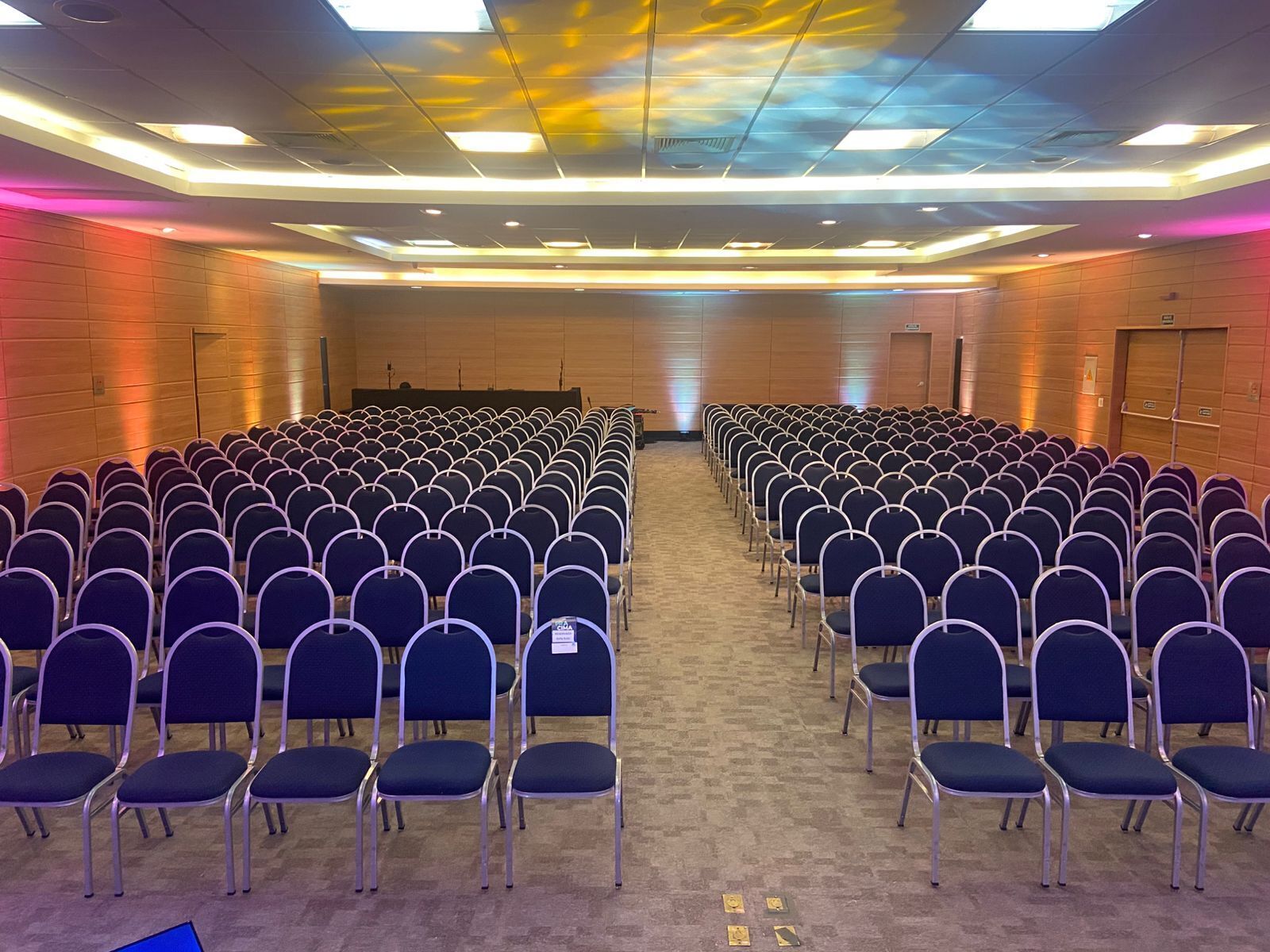A conference room with rows of chairs, a stage at the front, and a ceiling with recessed lighting.