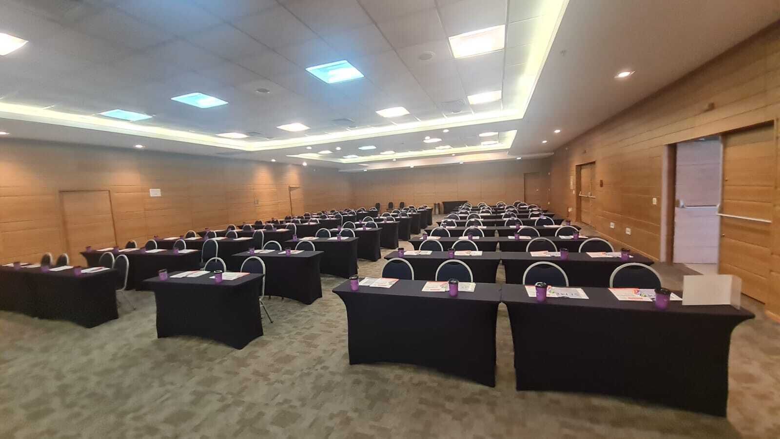 A conference room with rows of tables and chairs, equipped with nameplates and documents, in a room with wooden paneling and a carpeted floor.