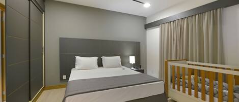 A modern hotel room with a large bed, a wooden crib, a nightstand, and a headboard.