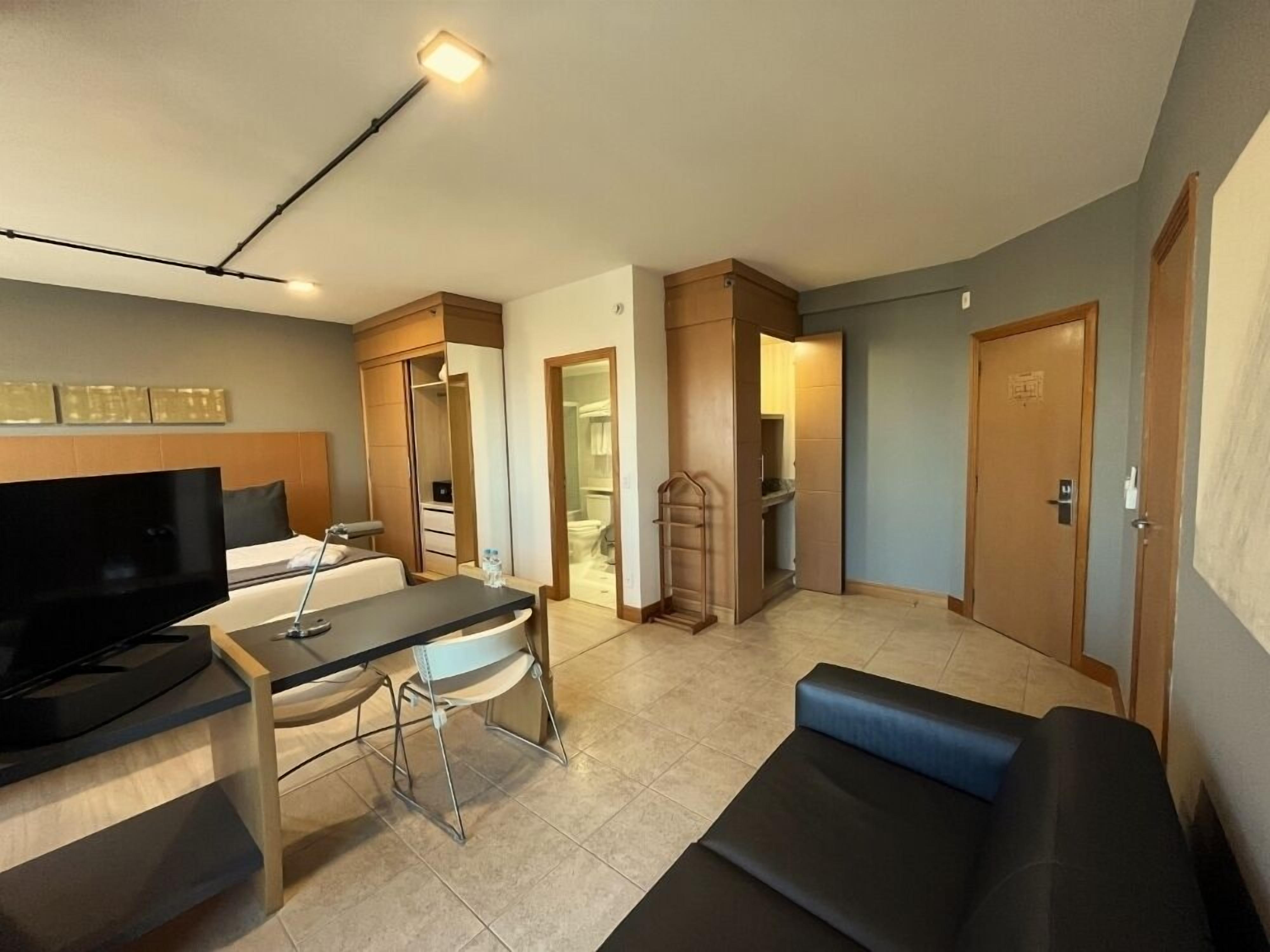 A modern hotel room with a flat-screen TV, a desk with a chair, a sofa, a wardrobe, and a bathroom visible through an open door.