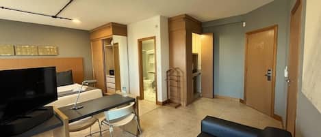 A modern hotel room with a flat-screen TV, a desk with a chair, a sofa, a wardrobe, and a bathroom visible through an open door.