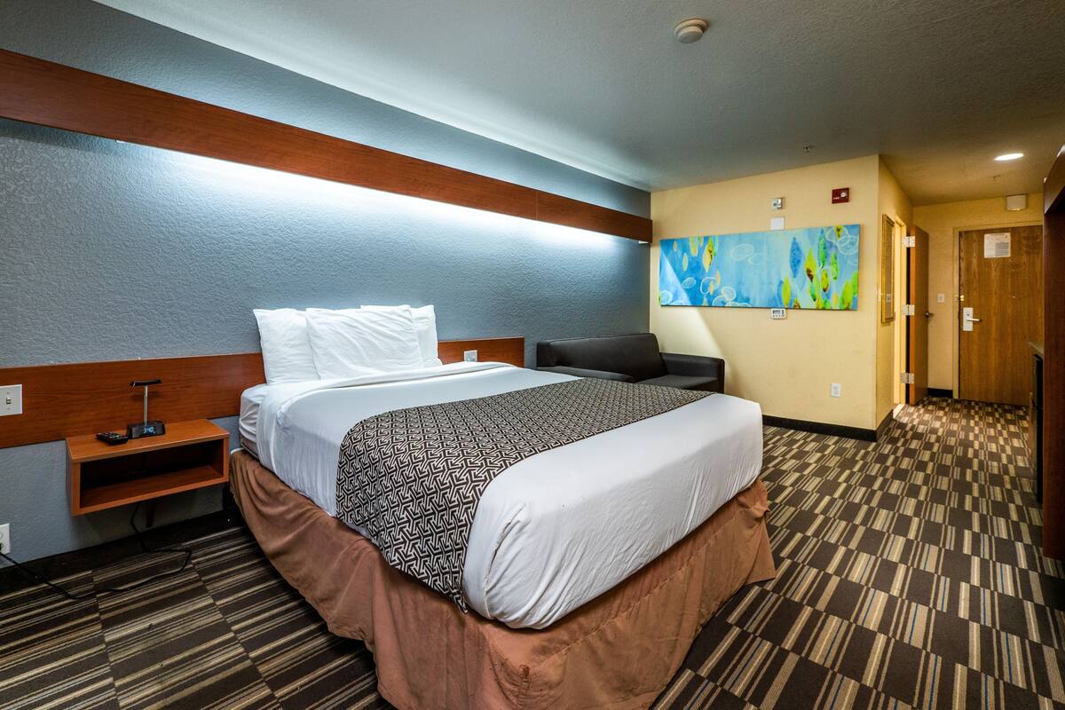 A hotel room with a bed, bedside table, lamp, and a painting on the wall.