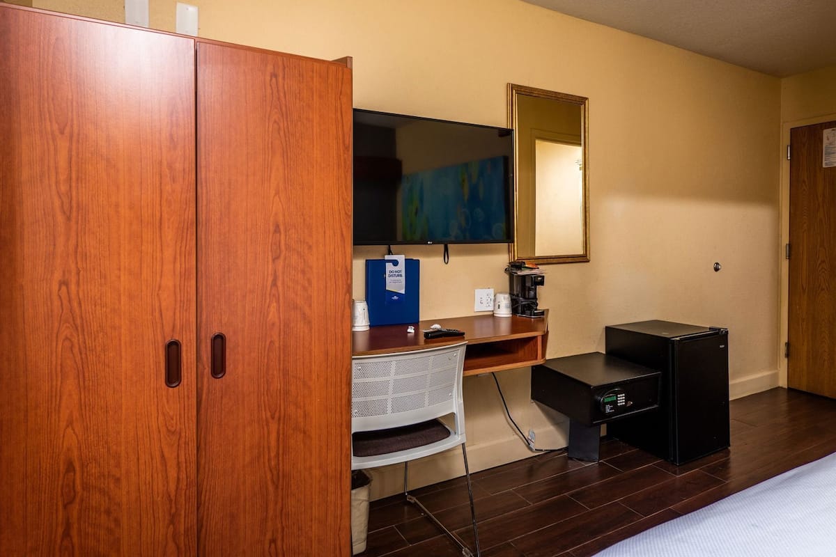 A hotel room with a wooden wardrobe, a desk with a computer, a coffee maker, and a small black speaker.