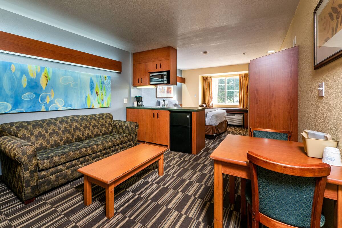 A compact hotel room with a sofa, a small dining table, a kitchenette, and a window with curtains.
