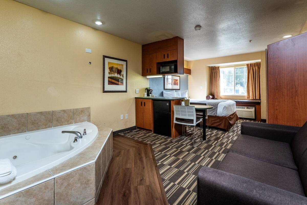 A compact hotel room with a large bathtub, a kitchenette, a dining area, and a window with curtains.