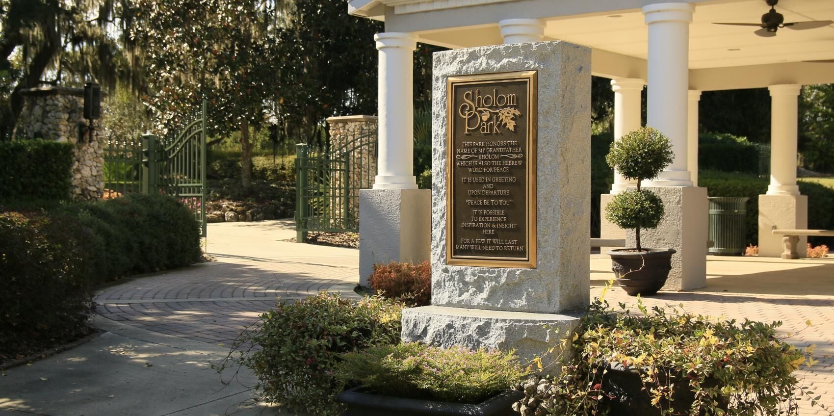 A stone monument with a plaque in a landscaped area with a covered walkway and potted plants.