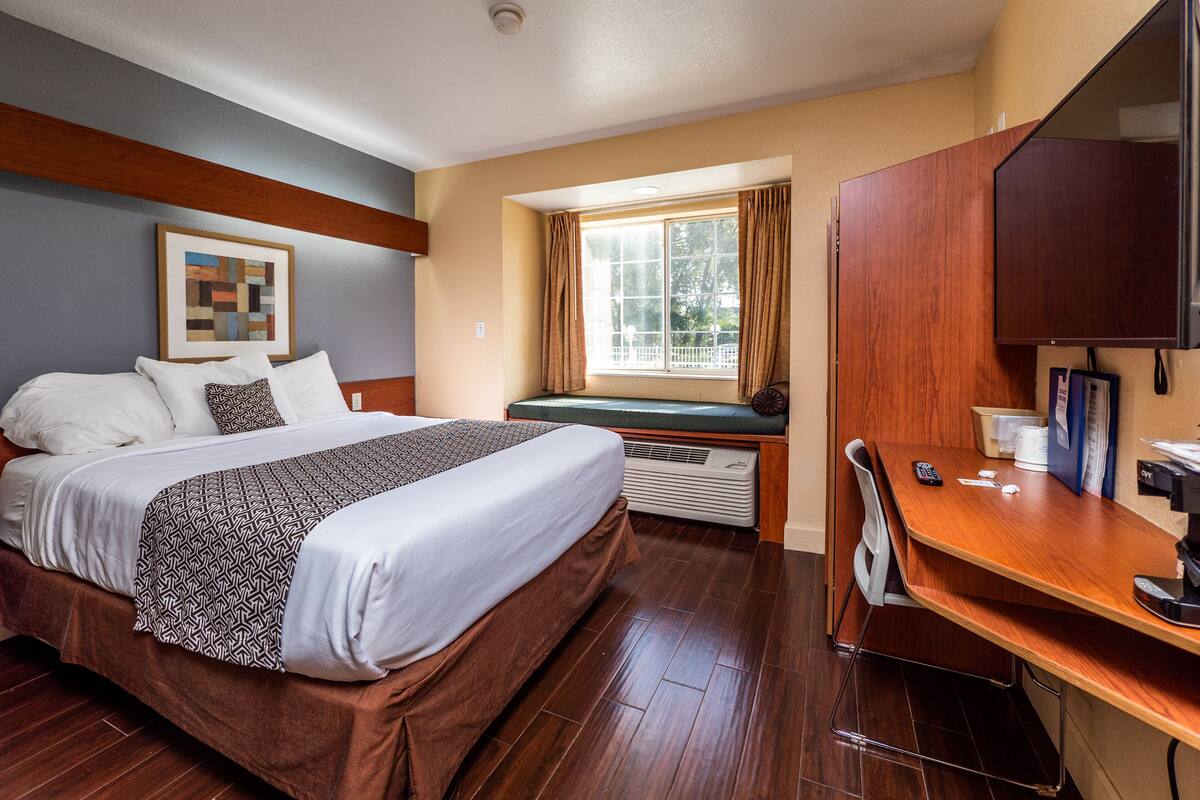 A hotel room with a bed, a desk, a chair, a window with curtains, a mounted TV, and a wall-mounted air conditioner.