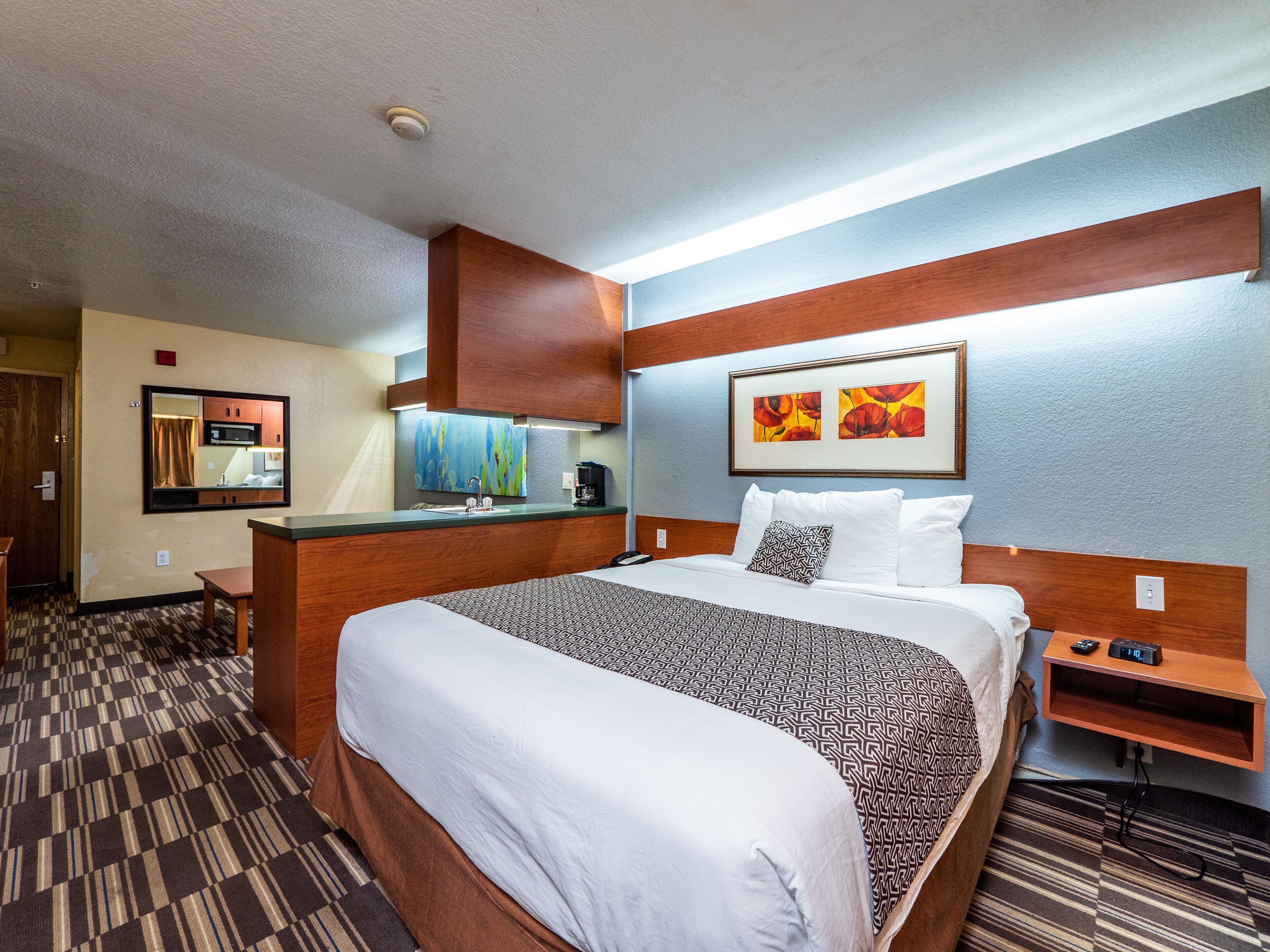 A hotel room with a large bed, a wooden headboard, a bedside table with a lamp, a microwave, and a painting on the wall.