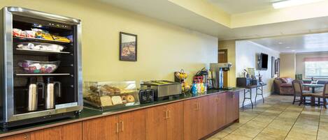 A kitchen area with a refrigerator, microwave, toaster, and coffee machine. There is a counter with various food items and a seating area with a table and chairs.