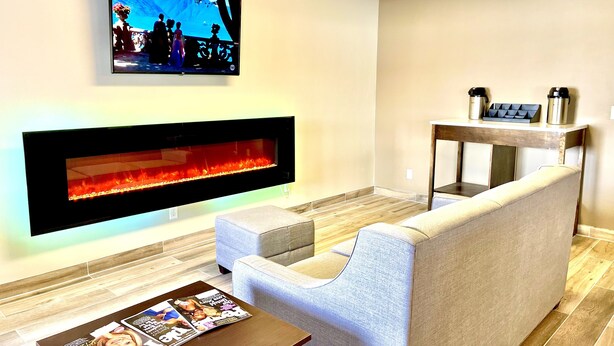 A modern living room with a flat-screen TV mounted on the wall, a built-in fireplace, a grey sofa, a coffee table with magazines, and a wooden side table with two coffee makers. - Econo Lodge Williston (Williston)