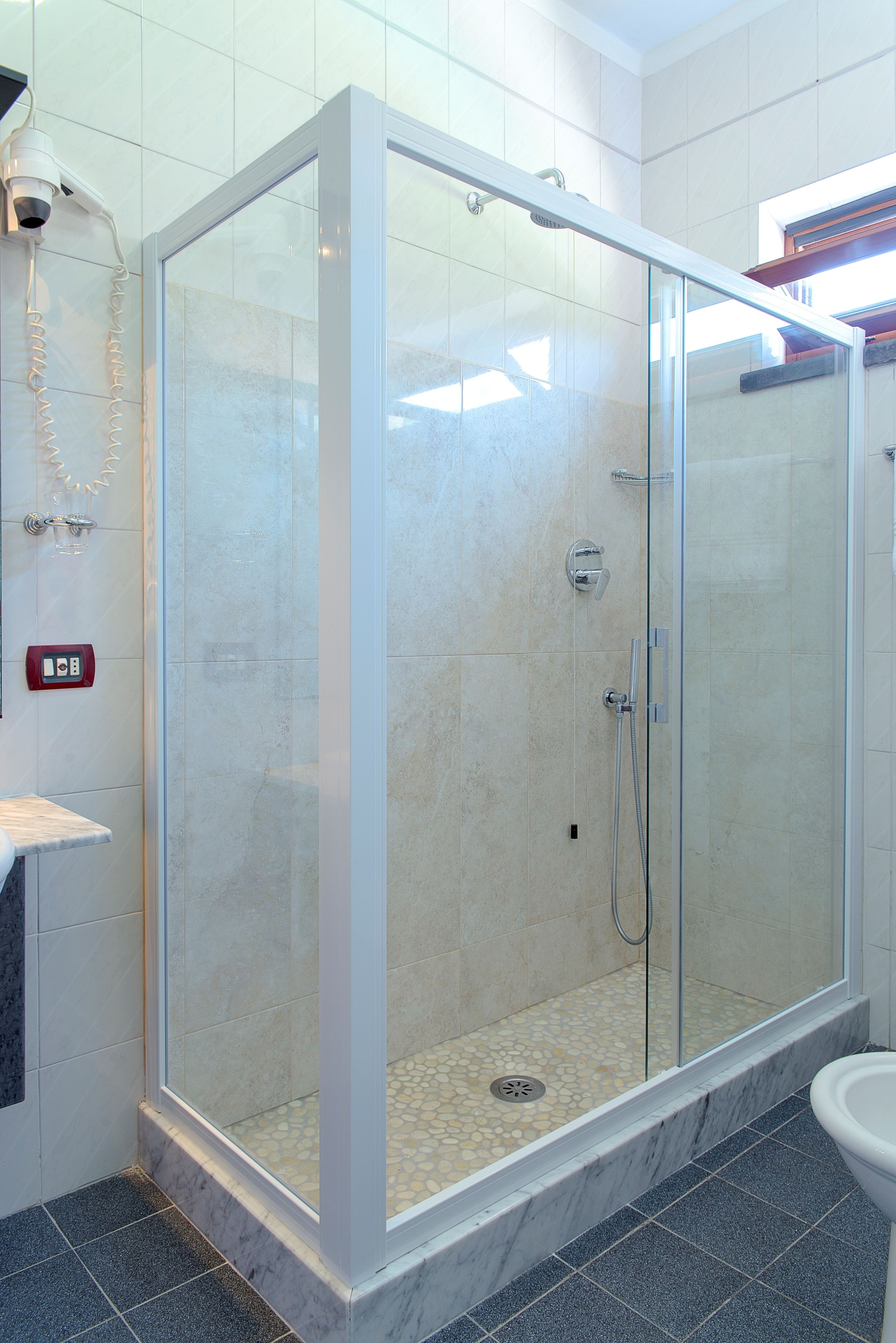 Ducha con paredes de vidrio, piso de baldosas y panel de control en la pared.