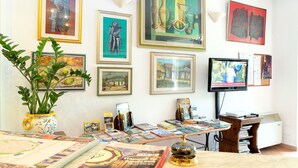 Een kamer met een tafel waarop boeken, tijdschriften, een potplant en meerdere ingelijste kunstwerken staan.