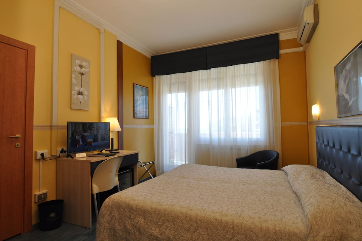 A hotel room with a bed, a desk with a television, a chair, and a window with curtains.