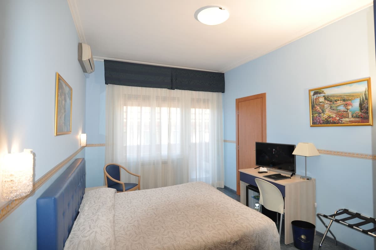 A hotel room with a bed, a desk with a computer, a chair, a window with sheer curtains, and a painting on the wall.