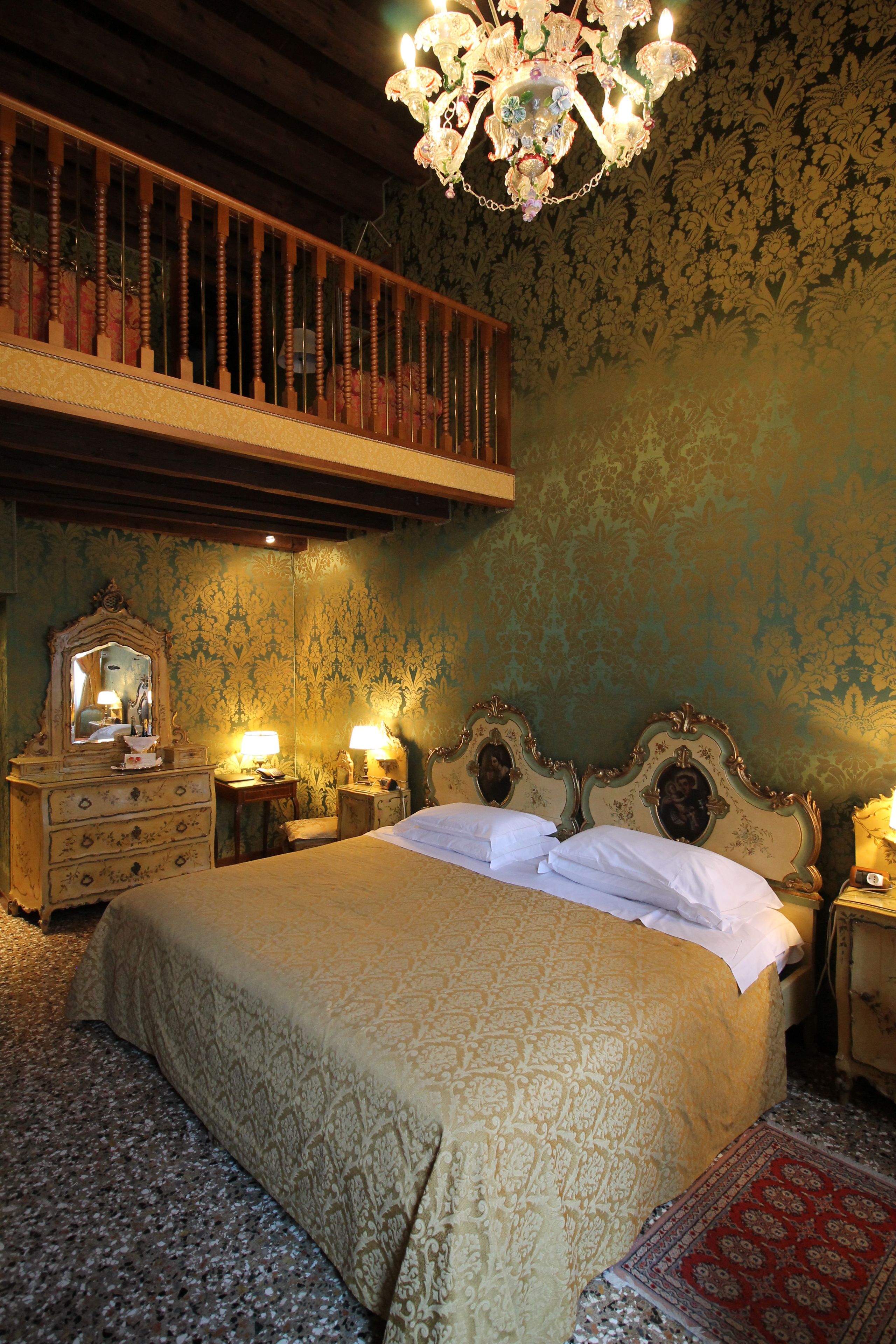 A bedroom with a large bed, ornate headboard, a chandelier, a wooden bench, and a patterned carpet.