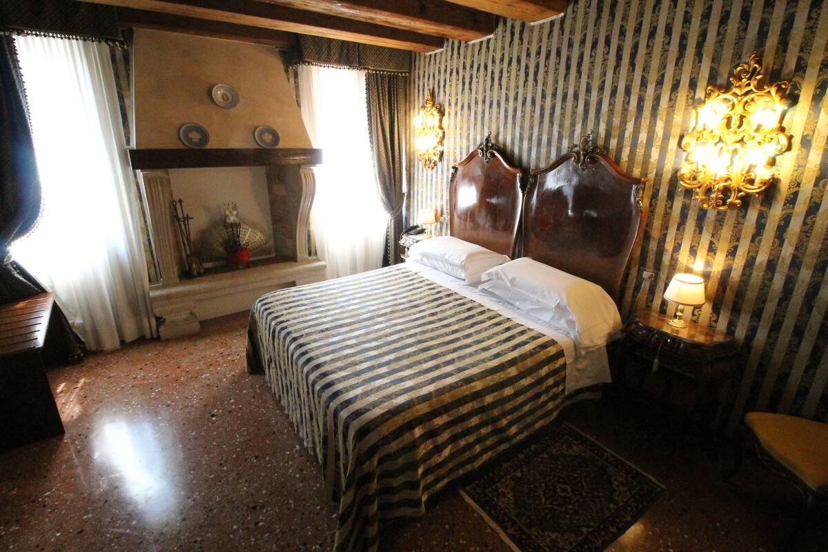 Een klassieke kamer met een groot bed, een houten hoofdbord, een nachtkastje met een lamp en behang met een patroon.
