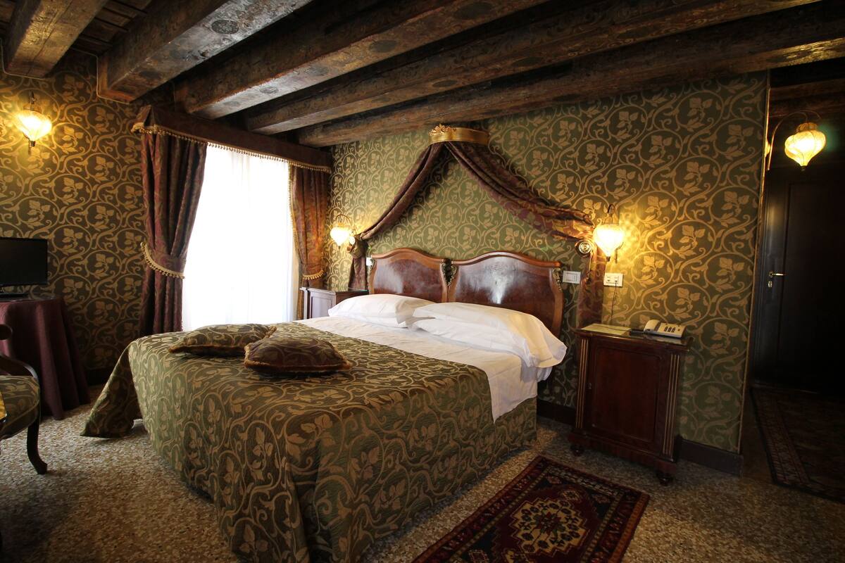 Een klassieke kamer met een hemelbed, behang met patroon en een houten plafond.