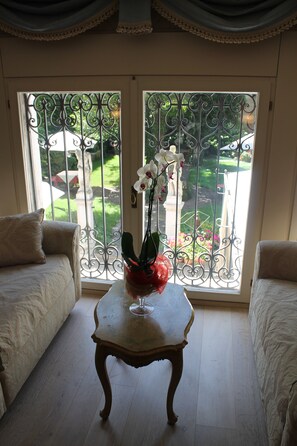 A room with a white sofa, a small wooden table with a vase of flowers, and a view through a metal gate to a garden. - Hotel Palazzo Abadessa (Venice)