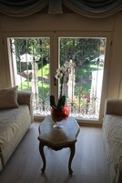 Ein Zimmer mit einem weißen Sofa, einem kleinen Holztisch mit einer Blumenvase und Blick durch ein Metallgitter auf einen Garten.