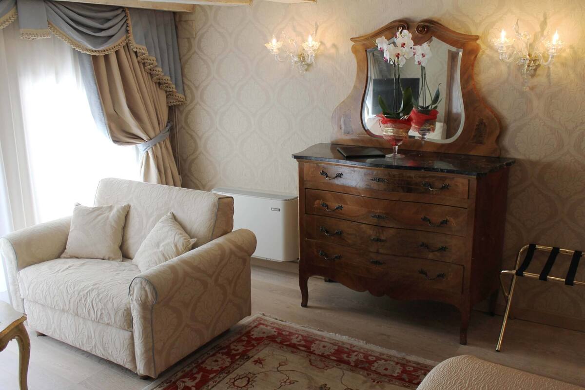 Een kamer met een beige fauteuil, een houten dressoir met spiegel en een rood tapijt met patroon.