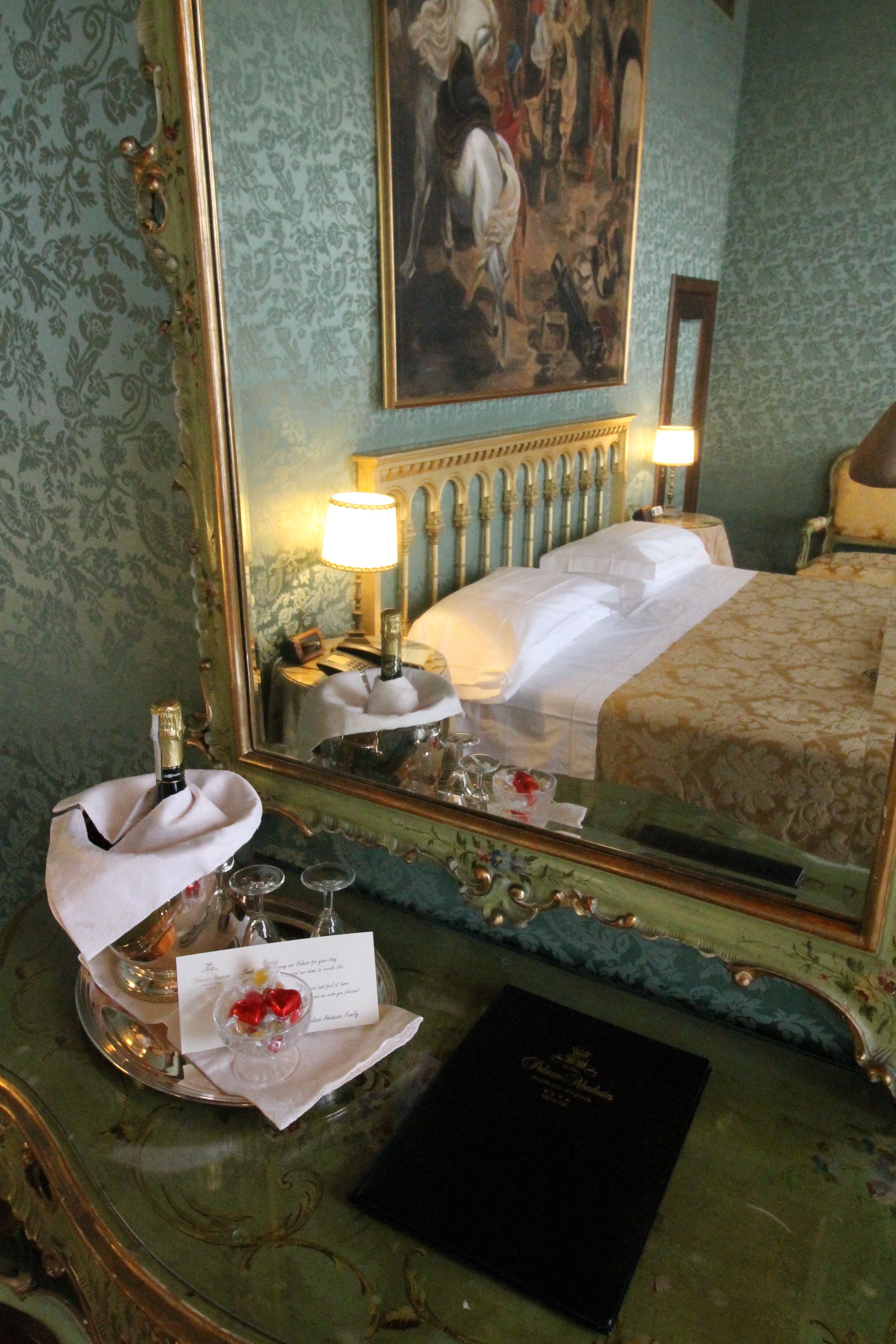 A hotel room with a bed, a nightstand, a mirror, a dresser, and a tray with a bottle and glasses.