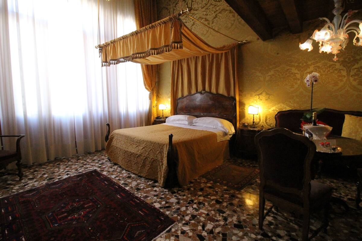 Een klassieke slaapkamer met een hemelbed, een zithoek met een stoel en een tafeltje, en een kroonluchter.