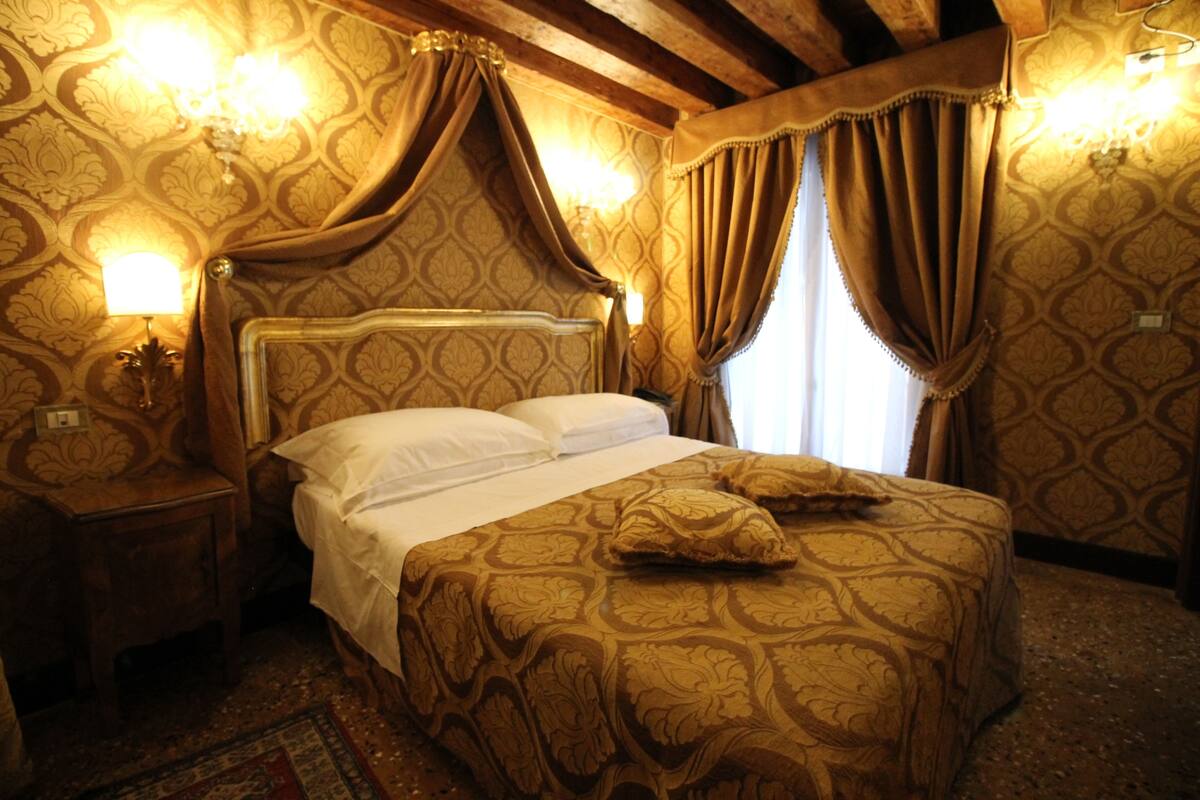 Een kamer met een groot bed, een versierd hoofdeinde en behang met een patroon. Er staan twee bedlampen en een nachtkastje.