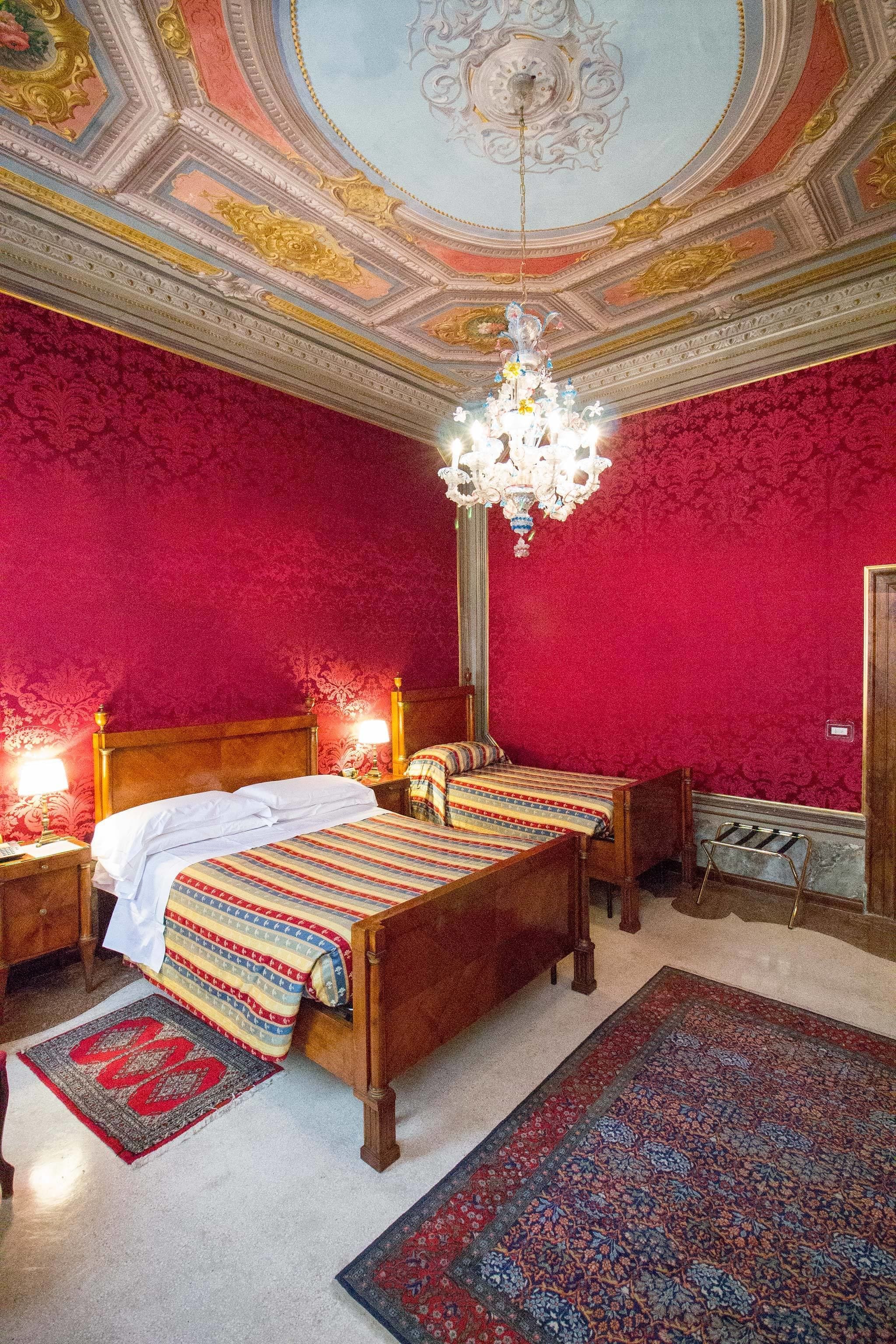 Une chambre avec un grand lit, deux tables de chevet, un lustre et un mur décoré d’un motif rouge.
