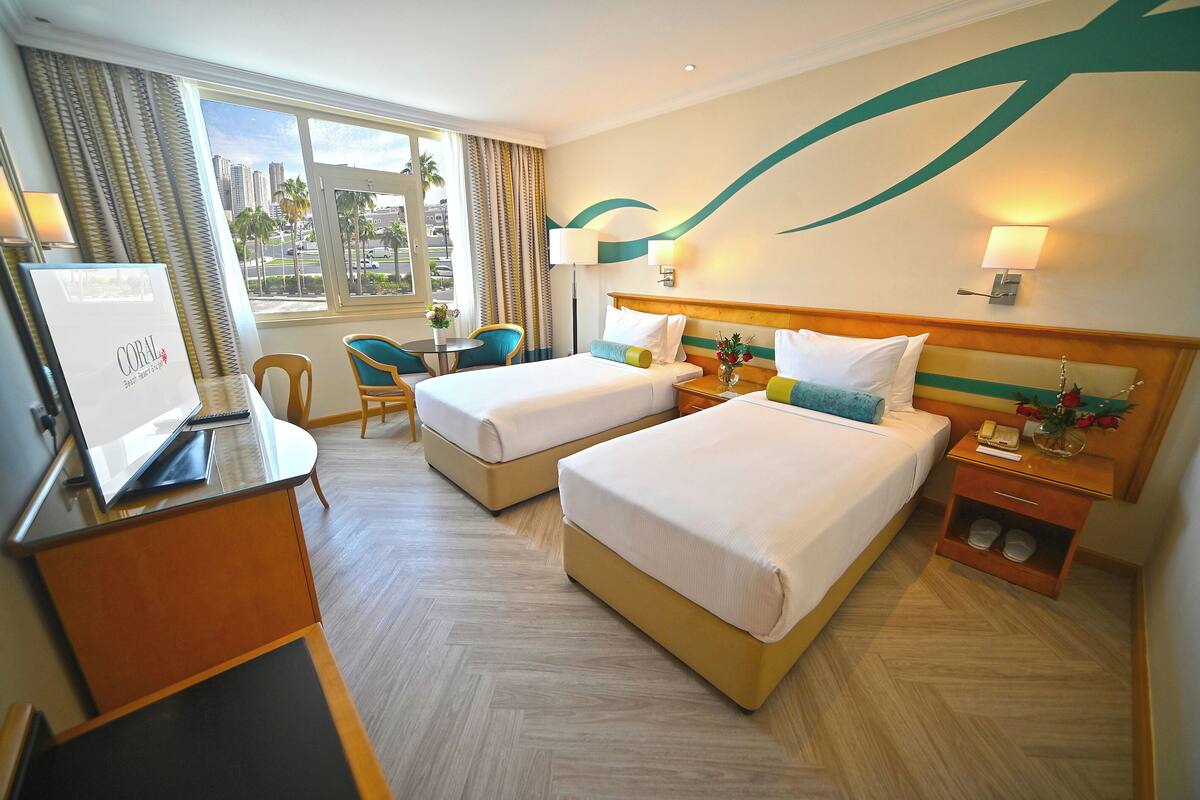 A hotel room with two beds, a desk with a computer, a chair, and a window with a view of palm trees and buildings.