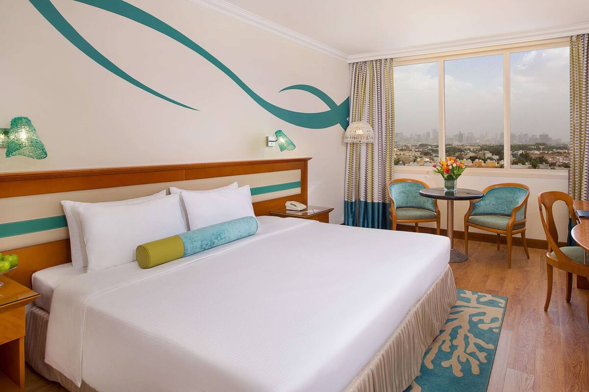 A hotel room with a large bed, a desk with a TV, a chair, a small table with a vase of flowers, and a view of a cityscape through the window.