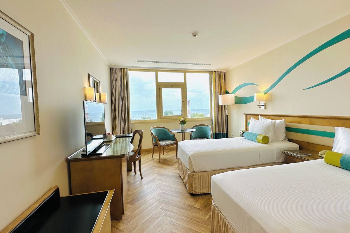 A hotel room with two beds, a desk, a chair, a small table, and a window with a view of the sea.