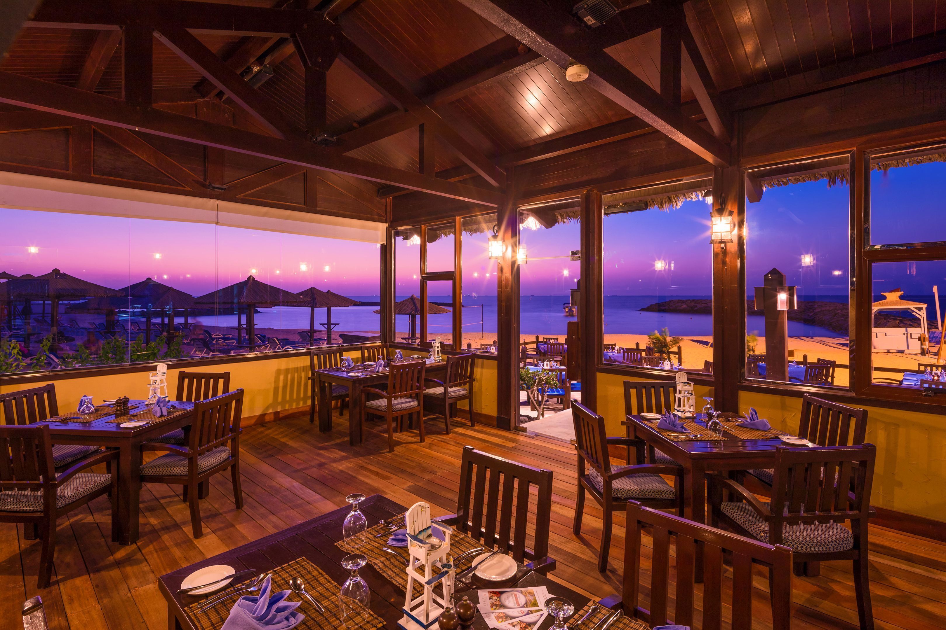 Un restaurante con mesas y sillas de madera, amplias ventanas con vista a la playa y sombrillas de paja.
