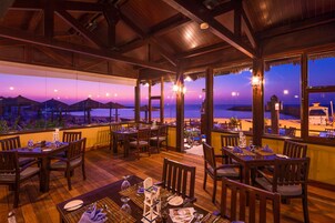 Un restaurante con mesas y sillas de madera, amplias ventanas con vista a la playa y sombrillas de paja.