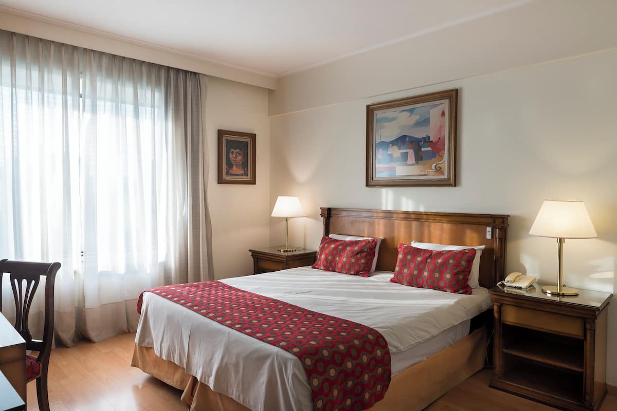 Quarto de hotel com uma cama grande, duas mesas de cabeceira com abajures, uma cadeira e um quadro na parede.