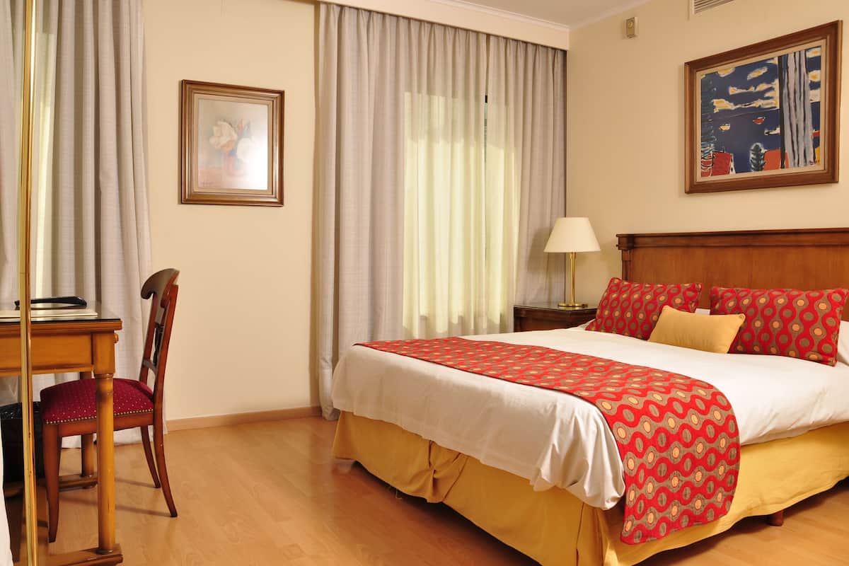 Quarto de hotel com cama, uma cadeira, uma escrivaninha, abajur e duas pinturas nas paredes.